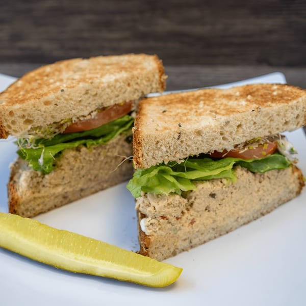 Tuna salad sandwich on toasted bread with lettuce, tomato, and sprouts, served with a pickle spear.