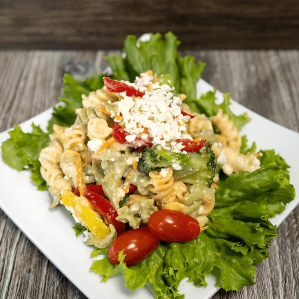 Pasta salad with rotini, bell peppers, broccoli, cherry tomatoes, and crumbled feta on lettuce.