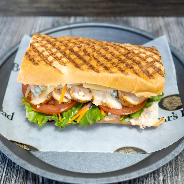 Grilled chicken panini with lettuce, tomato, shredded carrots, and blue cheese crumbles on a dark plate.