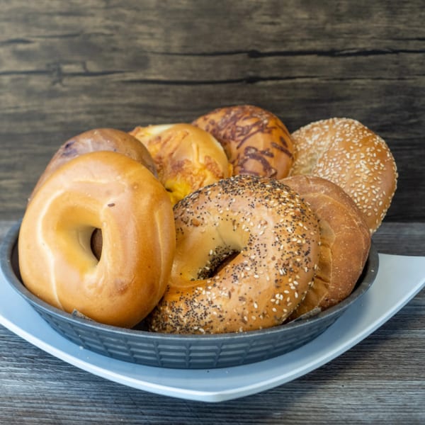 A basket of assorted bagels, including plain, sesame, poppy seed, and cheese, on a wooden surface.
